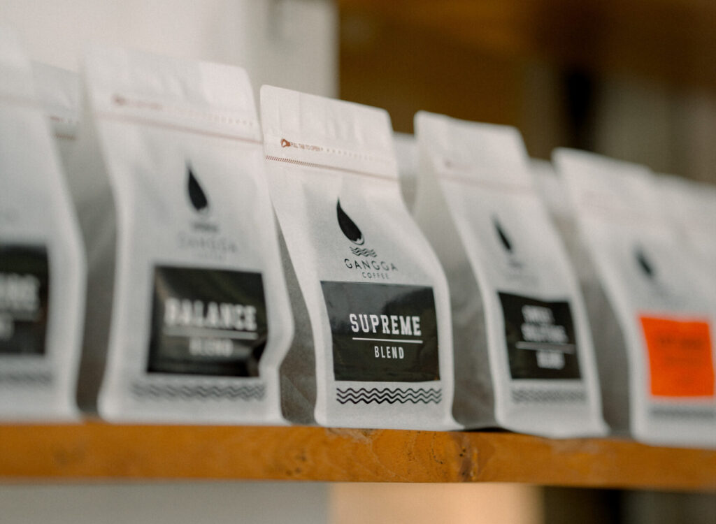 Gangga Coffee specialty coffee bags displayed on a wooden shelf, featuring various coffee blends in premium white packaging, artisan roasted coffee collection, specialty coffee product lineup, modern coffee shop interior, Indonesian coffee brand showcase, retail coffee display with selective focus and warm ambient background. 
